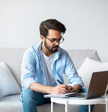 Online Education. Young Eastern Man Study With Laptop At Home And Taking Notes, Millennial Arab Guy In Eyeglasses Watching Online Webinar, Sitting On Couch With Computer, Enjoying Distance Learning