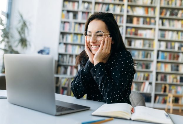 Joyful caucasian woman uses laptop. Happy female employee at online business meeting, dressed in stylish wear sit at the work desk, look at the laptop screen and smiles